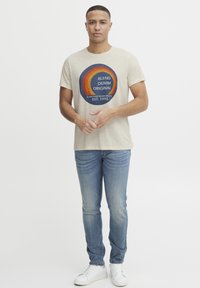 Camiseta de algodón color beige con gráfico circular multicolor que dice "BLEND DENIM ORIGINAL," combinada con jeans ajustados de color azul claro y zapatillas blancas.