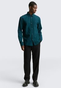 Tegen de diepblauwe achtergrond van een rustige oceaan, draagt een persoon een teal button-up overhemd met een kraag, lange mouwen en een subtiele textuur. Het overhemd is gecombineerd met zwarte broek en zwarte laarzen.