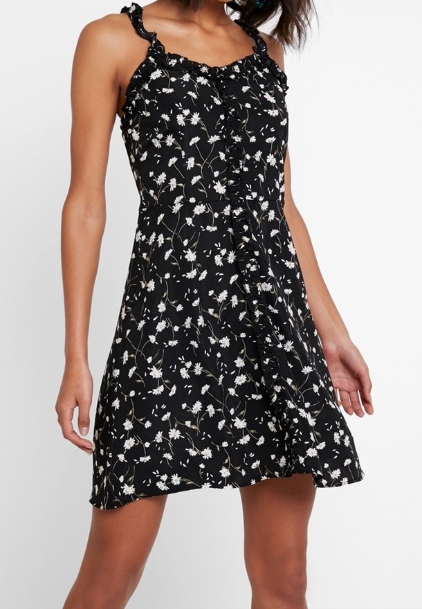 Femme portant une robe noire sans manches avec un motif floral blanc et des détails à volants sur le devant et les bretelles.
