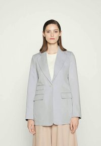 Blazer cinza claro com lapelas notched, dois bolsos frontais, fecho de botão único e tecido suave; combinado com uma camisa creme e uma saia bege.
