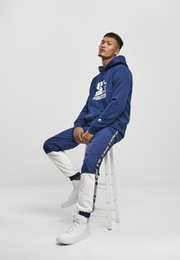 Tengerészeti kapucnis pulóver nagy fehér logóval, színek szerint tagolt nadrággal, amely fehér és tengerészkék részeket tartalmaz. Fehér magas szárú sportcipők.