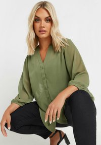 Blusa leggera color verde oliva con bottoni frontali, scollatura a V e maniche arricciate; abbinata a pantaloni neri e sandali.
