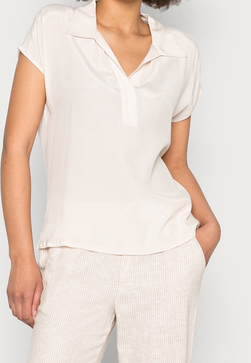 Femme en blouse crème à manches courtes avec col et pantalon beige clair rayé, une main dans la poche sur un fond uni.
