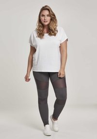 Weißes, eng anliegendes T-Shirt kombiniert mit dunkelgrauen Leggings, die Netzpaneele in einem geometrischen Muster aufweisen. Weiße Sneakers runden das Outfit ab.