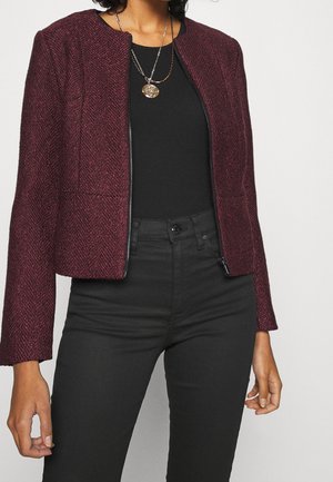 Femme portant une veste bordeaux texturée à zip sur un top noir, un pantalon noir taille haute et des colliers avec pendentifs dorés.