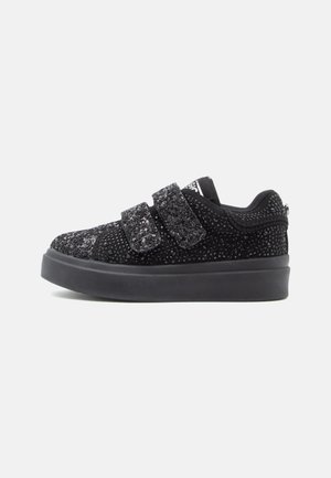 Zapatilla negra de corte bajo con suela gruesa, cubierta de pequeños remaches brillantes, que cuenta con dos amplias tiras de velcro para el cierre.