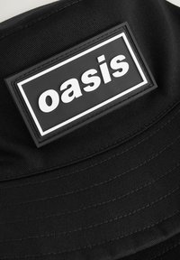 Musta ämpärilakki, joka on valmistettu kestävästä kankaasta, ja jossa on teksturoitu pinta sekä suorakulmainen kangas, jossa on sana "oasis" paksuilla valkoisilla kirjaimilla.