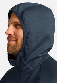 Giacca con cappuccio in tessuto blu scuro con una texture liscia. Presenta un cappuccio aderente e una chiusura frontale con zip, che mette in evidenza il collo.