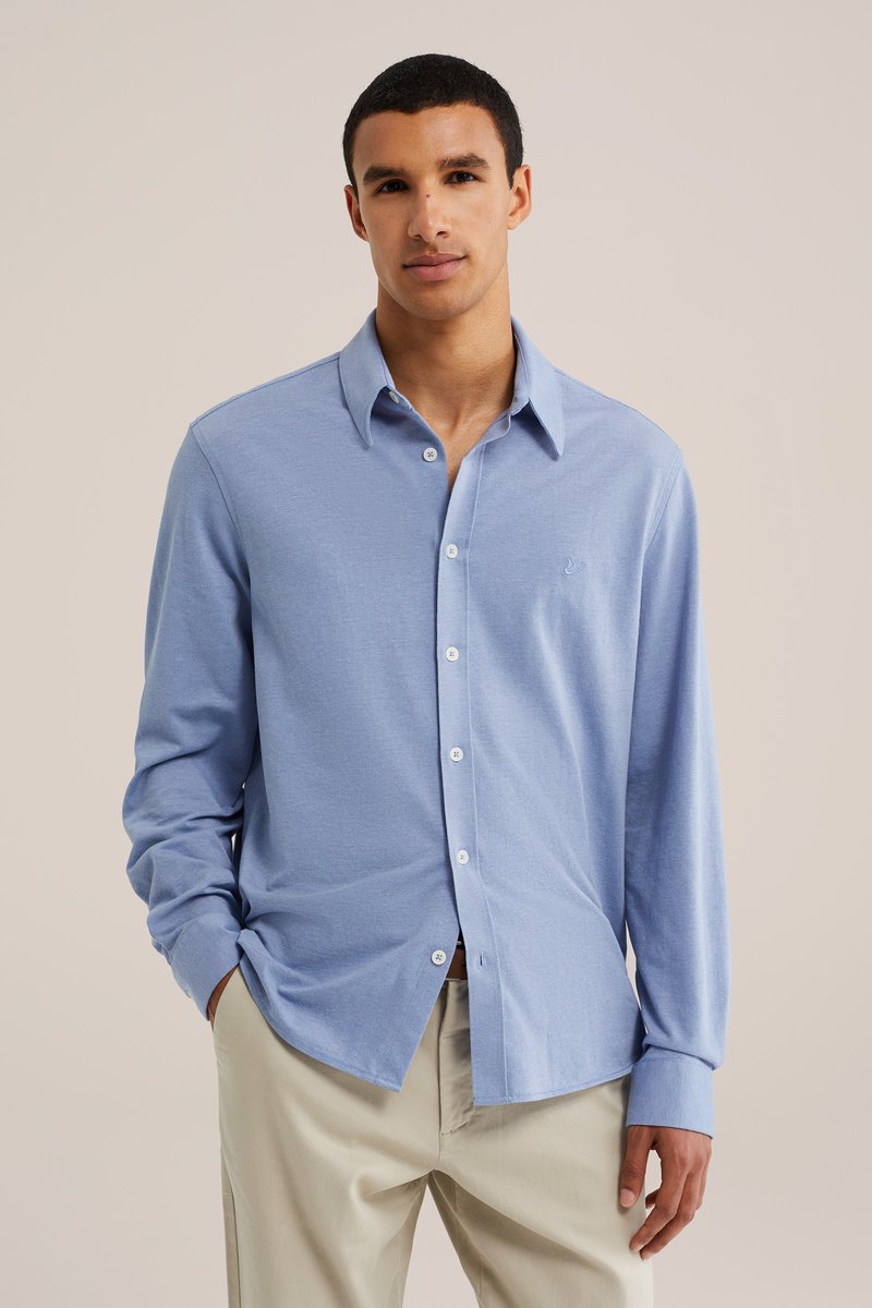 Jeune homme portant une chemise bleu clair à manches longues boutonnée et un pantalon beige, debout les mains dans les poches devant un fond uni.