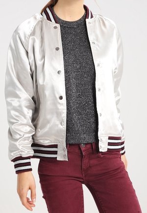 Femme portant une veste bomber argentée brillante avec des poignets rayés par-dessus un pull gris foncé et un pantalon bordeaux.