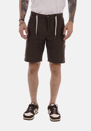 Uomo che indossa pantaloncini marrone scuro con cordini bianchi, scarpe da ginnastica bianche e nere e una camicia bianca, in piedi con le braccia tatuate visibili.