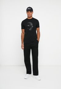 Hombre vestido con camiseta gráfica negra, pantalones cargo negros, gorra negra y zapatillas blancas de pie frente a un fondo blanco.