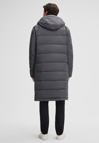 Grijze gewatteerde lange jas met een capuchon, met horizontale gequilte secties en een gladde textuur. Gecombineerd met zwarte broek en witte sneakers.