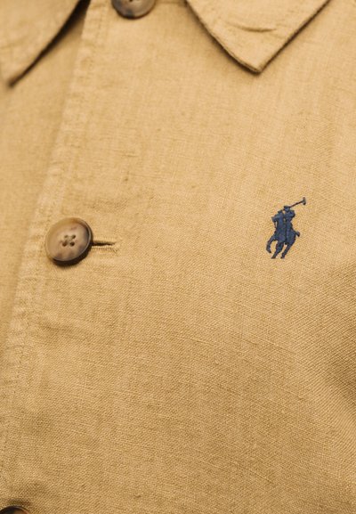 Κοντινή λήψη μπεζ λινό πουκάμισο με καφέ κουμπιά και μικρό σκούρο μπλε κεντητό παίκτη πόλο πάνω σε άλογο.