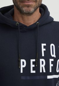 Kék kapucnis pulóver húzózsinórral és ezüst díszítéssel; nagy fehér felirattal rendelkezik, ami "FQ PERF"-ot olvasható. Puha anyag, laza kialakítás.