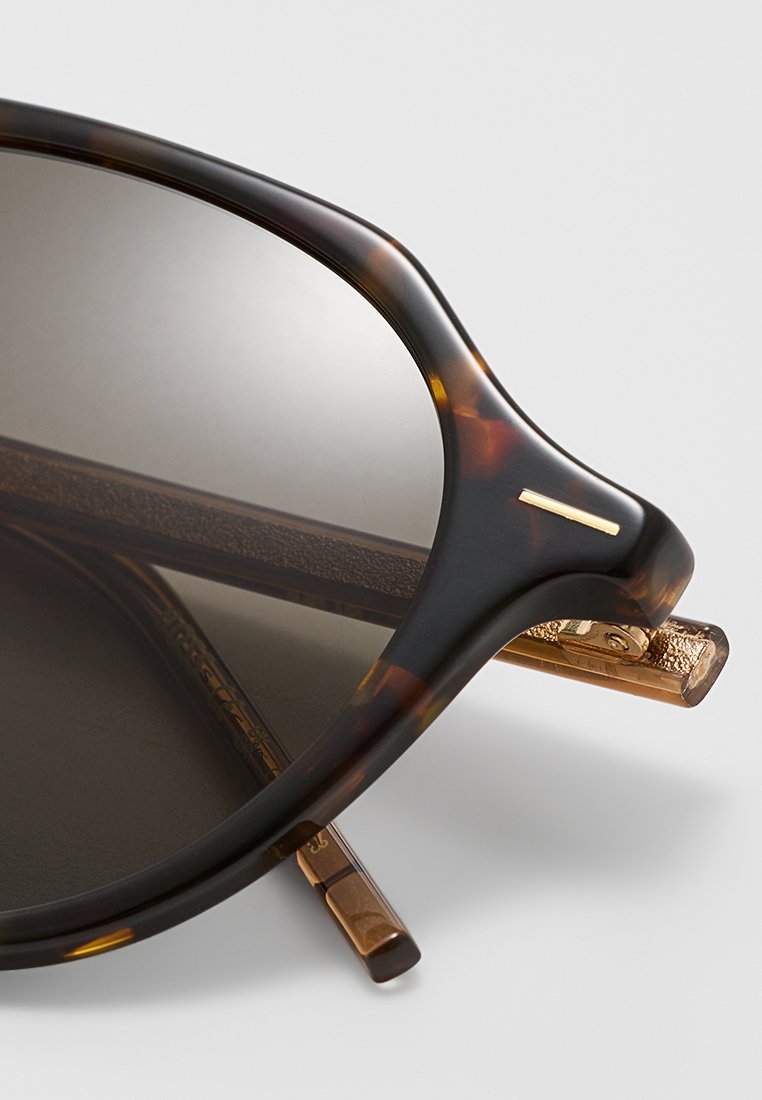 Lunettes de soleil en écaille de tortue avec des verres foncés, mettant en avant des accents en métal doré et un design de branches texturé. Forme ronde et bulbeuse.