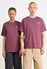 Deux individus se tenant côte à côte, portant des t-shirts en coton bordeaux à manches courtes et amples, avec un petit logo blanc sur la poitrine.