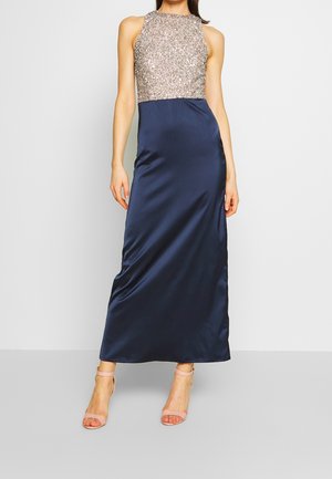 Kobieta w granatowej, satynowej sukni bez rękawów, sięgającej do podłogi, z wysokim dekoltem, brokatowym, srebrnym zdobionym górnym elementem i cielistymi szpilkami z paseczkiem wokół kostki.