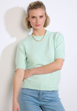 Kobieta o krótkich blond włosach, ubrana w jasnzielony sweter z krótkim rękawem, z złotym łańcuszkiem na szyi i jasnoniebieskie dżinsy, na białym tle.