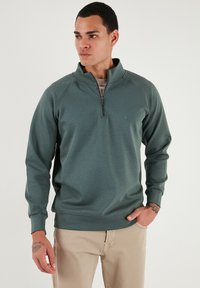 Sudadera verde con cremallera de cuarto, con una superficie texturizada, puños acanalados y un pequeño logo en el pecho. Combinada con pantalones de un beige claro.