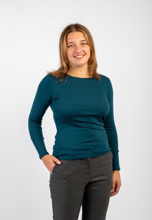 Camicia a maniche lunghe color turchese con scollo rotondo, taglio aderente, abbinata a pantaloni grigi scuri, tessuto liscio e senza motivi visibili.