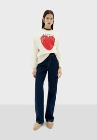 Crème lange mouwen shirt met rode grafiek en "ARTE" tekst, gecombineerd met blauwe high-waisted jeans en beige schoeisel. Eenvoudig ontwerp en casual pasvorm.