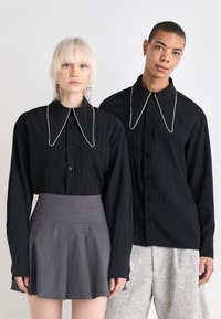 Camisas pretas de botão com grandes golas, apresentando detalhes semelhantes a pérolas. Uma pessoa usa uma saia plissada cinza, enquanto a outra veste calças prateadas cobertas de lantejoulas.