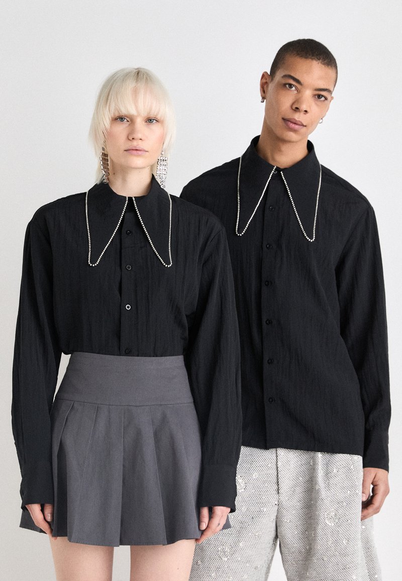 Camisas pretas de botão com grandes golas, apresentando detalhes semelhantes a pérolas. Uma pessoa usa uma saia plissada cinza, enquanto a outra veste calças prateadas cobertas de lantejoulas.
