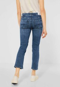 Jean en denim bleu avec une jambe droite, des poches arrière et une taille mi-haute. Porté avec un haut léger et des chaussures à plateforme beiges.