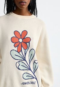 Pull en maille crème avec un grand motif de fleur rouge et de feuilles vertes. Texte « SANTA CRUZ » brodé près du bas en bleu marine.