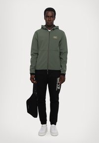 Felpa verde con cerniera e dettagli neri, abbinata a pantaloni neri con testo del logo bianco, sneakers bianche e una piccola borsa nera.