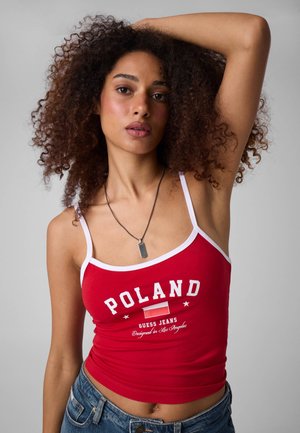 Frau mit lockigem Haar, die ein rotes Tanktop mit dem Text "POLAND" und blaue Jeans trägt, hebt einen Arm hinter den Kopf vor einem einfarbigen Hintergrund.