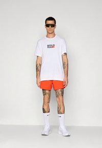 Vit t-shirt med korta ärmar och "BERLIN RUN"-tryck, kombinerad med klarröda shorts. Inkluderar solglasögon, vita sneakers och strumpor som går upp till vaden.