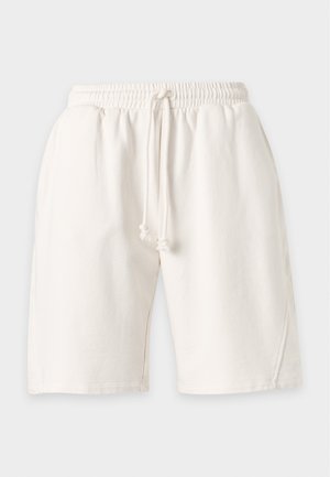 Γυναίκα στέκεται με το χέρι στη μέση, φοράει λευκό αμάνικο τοπ, λευκά σορτς με κορδόνι μέχρι το γόνατο και μαύρα σανδάλια με τακούνι.