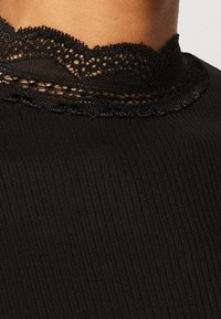 Zwarte top met een getextureerde geribbelde stof. De kraag is afgewerkt met een gedetailleerd kanten randje met een golvende rand.