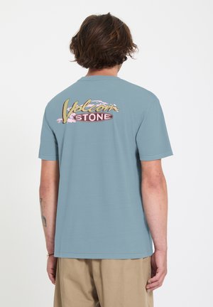 Homme aux cheveux mi-longs portant un T-shirt Volcom Stone bleu et un pantalon beige, vu de dos sur un fond uni.