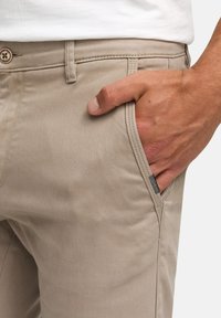 Beige katoenen broek met een gestructureerde afwerking, voorzien van een voorzak en een gestikte tailleband. Klein merklabel zichtbaar aan de zijkant.
