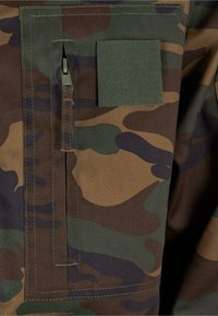 Zöld, barna és fekete árnyalatú kamuflázs anyag. Jellemzője egy cipzáros zseb és egy tépőzáras folt a strukturált dizájnon. Texturált felület.