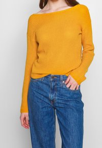 Mosterdgele off-shoulder trui met geribbelde textuur, gecombineerd met high-waisted blauwe jeans. Voorzien van lange mouwen en een aansluitend ontwerp.