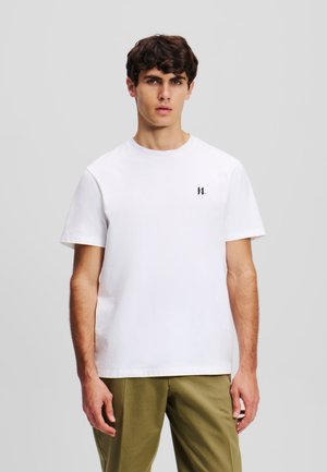 Jonge man met kort, krullend haar draagt een wit T-shirt met een subtiel logo, gecombineerd met olijfgroene broek, tegen een neutrale grijze achtergrond.