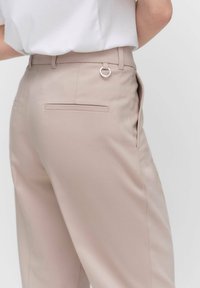 Beige, muotoon ommellut housut nähtynä takaa, joissa on metallirengasdetalji vyötäröllä ja kaksi takataskua tasaisella muotoilulla.