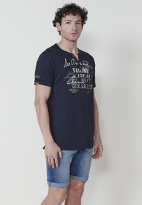 Tumši zils kokvilnas T-krekls ar īsām piedurknēm, apaļu kakla izgriezumu un kontrastējošām baltām tekstu grafiskām detaļām. Apvienots ar džinsu šortiem un sarullētām piedurknēm.