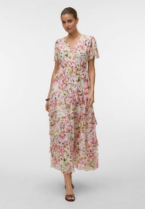 Femme portant une robe maxi à volants fleurie rose et blanche avec manches courtes et col en V, associée à des talons bruns à brides et un bracelet.