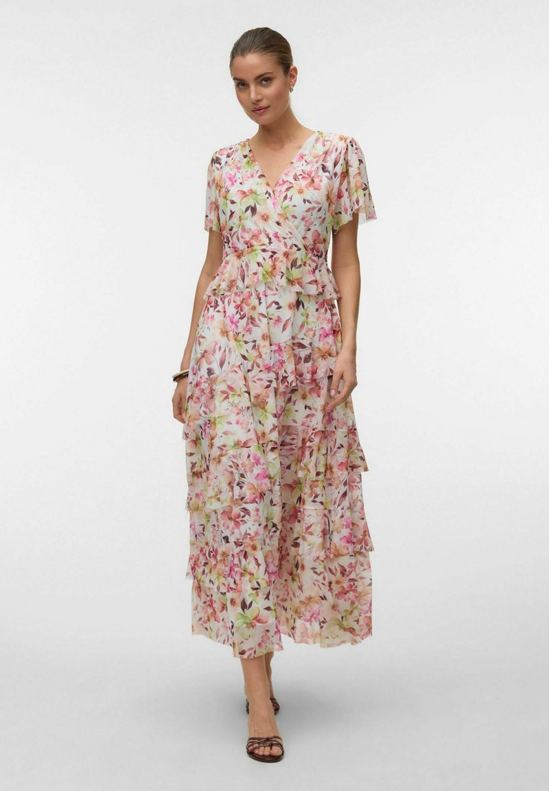 Donna che indossa un vestito lungo a balze floreale rosa e bianco con maniche corte e scollo a V, abbinato a tacchi con cinturini marroni e un braccialetto.