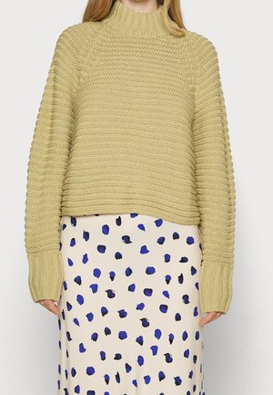 Femme portant un pull à col roulé beige texturé et une jupe blanche avec des taches irrégulières bleues et noires, debout devant un fond uni.