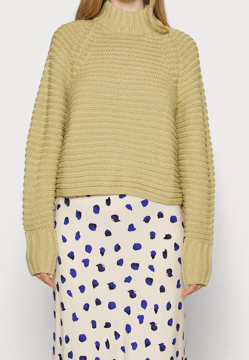 Femme portant un pull à col roulé beige texturé et une jupe blanche avec des taches irrégulières bleues et noires, debout devant un fond uni.