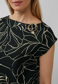 Zwarte top met wit bloemenpatroon, gestructureerde stof en ronde halslijn. Voorzien van een ronde gouden accentspeld op de schouder.