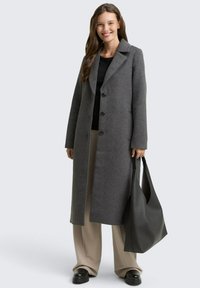 Vrouw in een lange grijze knopenjas, zwarte top, beige wijde broek, zwarte schoenen en met een grote zwarte handtas.