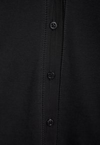Cardigan noir avec un devant boutonné, texture tricot lisse et légère brillance, avec six boutons noirs ronds alignés verticalement.