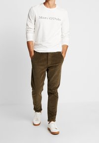 Bílá dlouhá košile s nápisem "Marc O'Polo", olivově zelené kalhoty a bílé tenisky s hnědými podrážkami, ukazující neformální outfit.